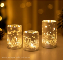 Cargar imagen en el visor de la galería, Set of three silver frosted glass candle holders glowing with winter village designs, reindeer, and snowflakes against a warm golden background.