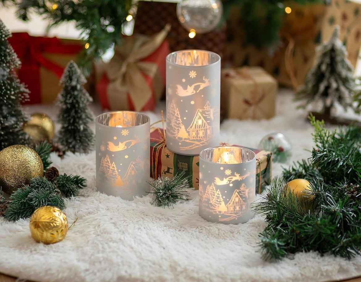 Silver frosted winter village candle holders styled with Christmas greenery, gold ornaments, and wrapped gifts under a decorated tree.
