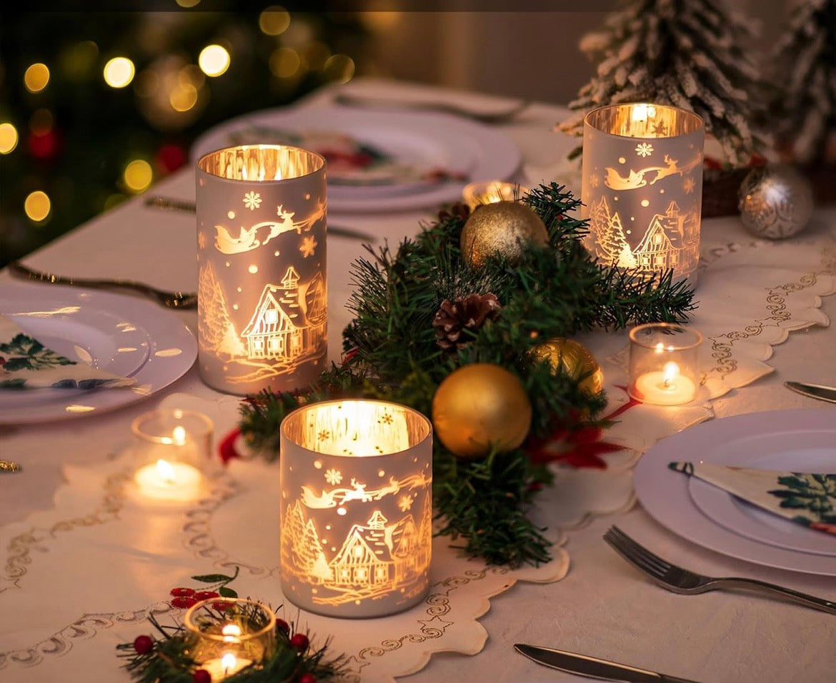 Winter village frosted glass candle holders used as a Christmas table centerpiece surrounded by plates, greenery, and warm candlelight.